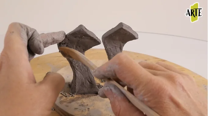Manos trabajando dos formas de pasta de modelar con un palillo sobre una base giratoria