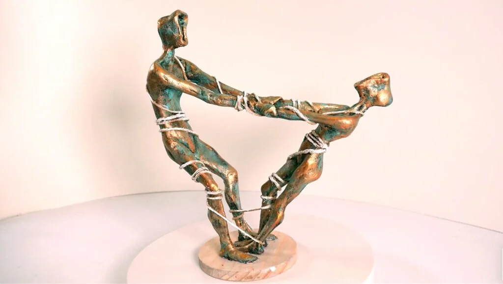 Dos figuras estilizadas entrelazadas, modeladas y pintadas con efecto bronce y pátina verdosa