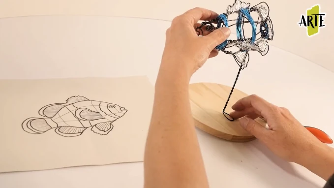 Manos trabajando una figura de pez en alambre sobre una base de madera junto a su dibujo